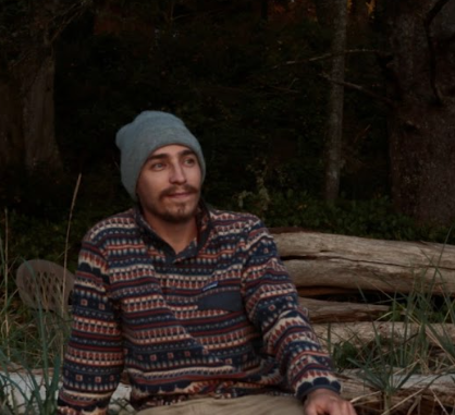 Ari Blaff seated outdoors on a beach, wearing a light blue beanie and a patterned fleece pullover